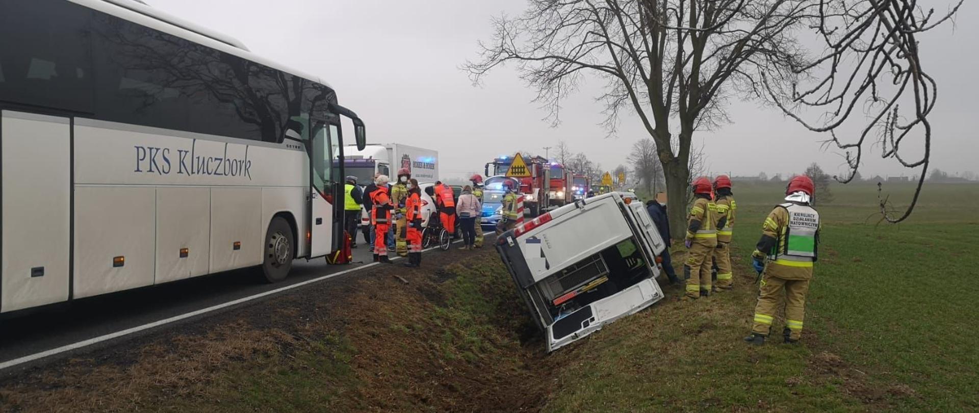 Zdjęcie przedstawia przewrócony na bok samochód typu bus w rowie przy drodze krajowej nr 11. Na zdjęciu widoczni również strażacy w ubraniu specjalnym koloru piaskowego i czerwonym hełmie na głowie, członkowie zespołów ratownictwa medycznego oraz policjanci. Po lewej stronie autobus PKS Kluczbork.