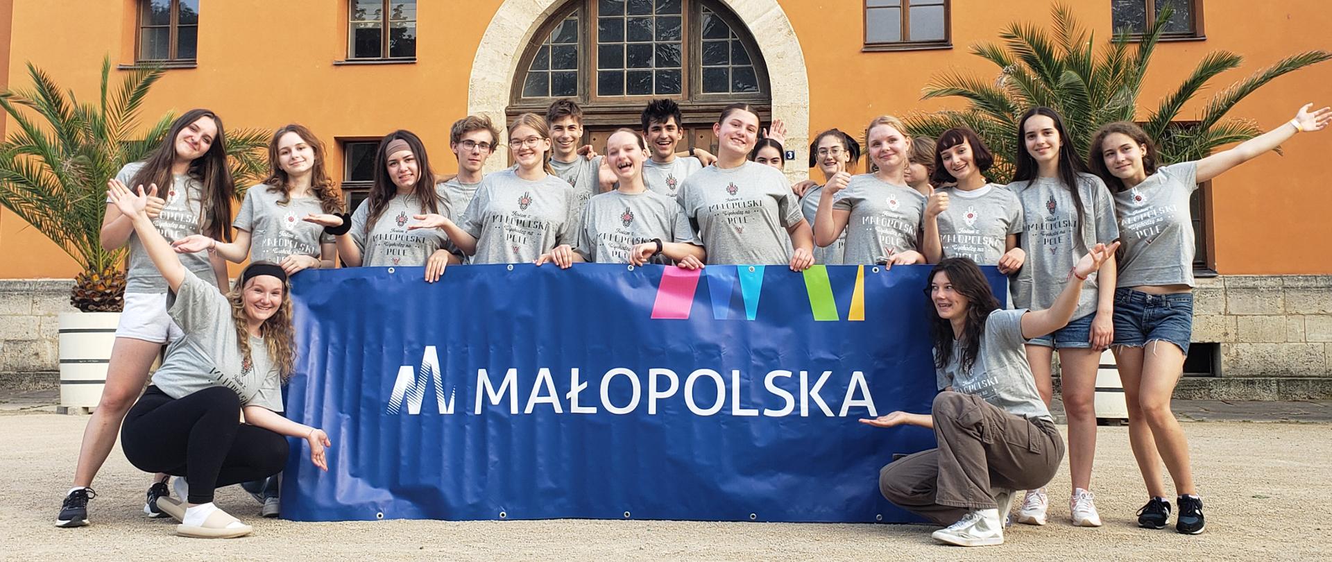 Zdjęcie grupowe uczniów będących na wyjeździe w Niemczech. W tle budynek z wieloma oknami ora dwoma palmami. Uczniowie pozują do zdjęcia trzymając baner współorganizatora Urząd Marszałkowski. Uczniowie ubrani są w jednakowe szare koszulki z logo "Małopolska"