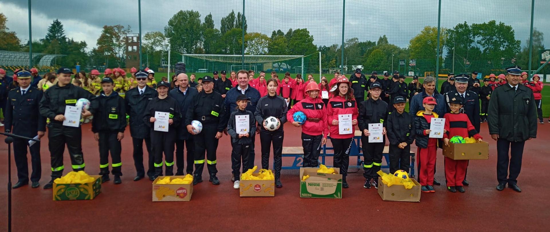 Powiatowe Zawody Sportowo Pożarnicze Młodzieżowych Drużyn Pożarniczych OSP 2022 - - na zdjęciu druhowie OSP w stoją szeregu z dyplomami w rękach z strażakami w mundurach wyjściowych podczas zbiórki na tartanie na boisku