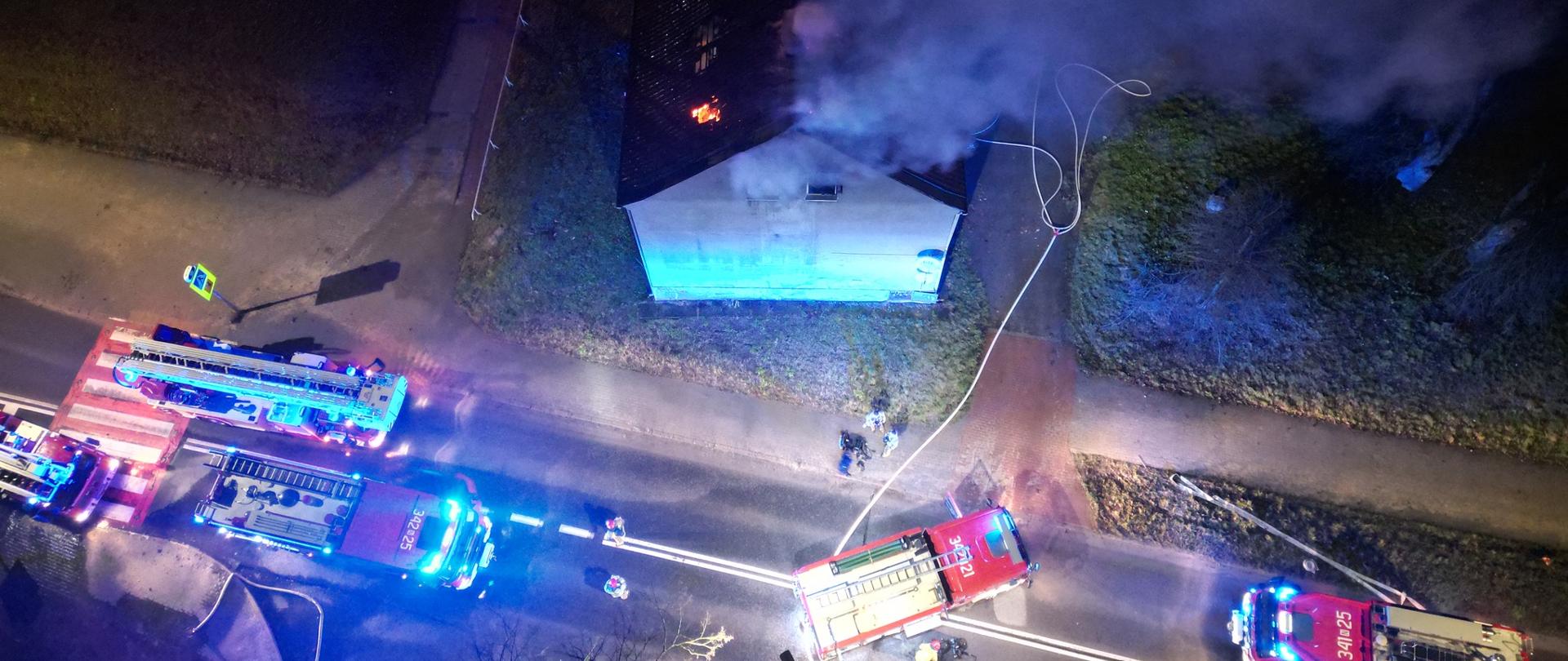 Widok z góry na palący się dach pustostanu. Obok widoczne pojazdy pożarnicze i sylwetki strażaków. Dookoła znajdują się kłęby dymu.