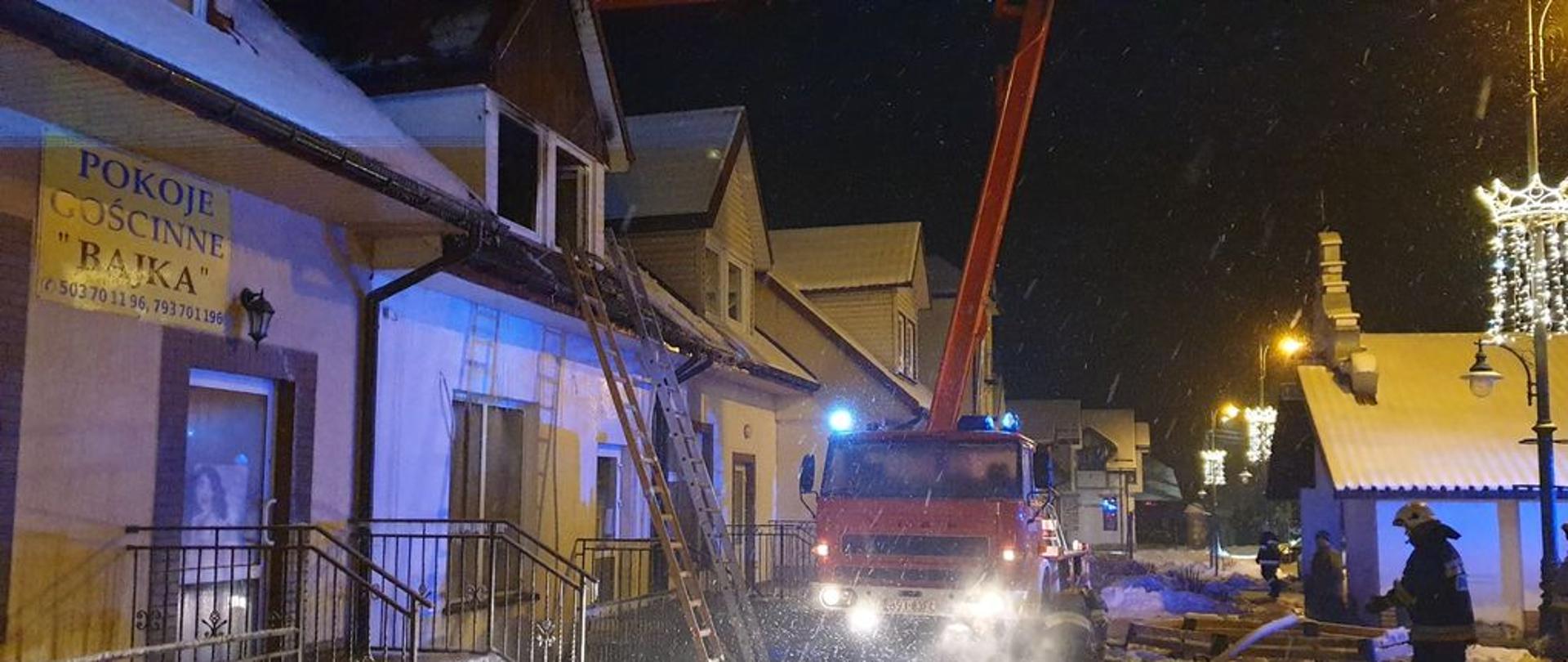 Palący się budynek parterowy kamienicy z poddaszem użytkowym. Przy budynku rozstawiony podnośnik strażacki oraz strażacy wykonujący czynności ratownicze.