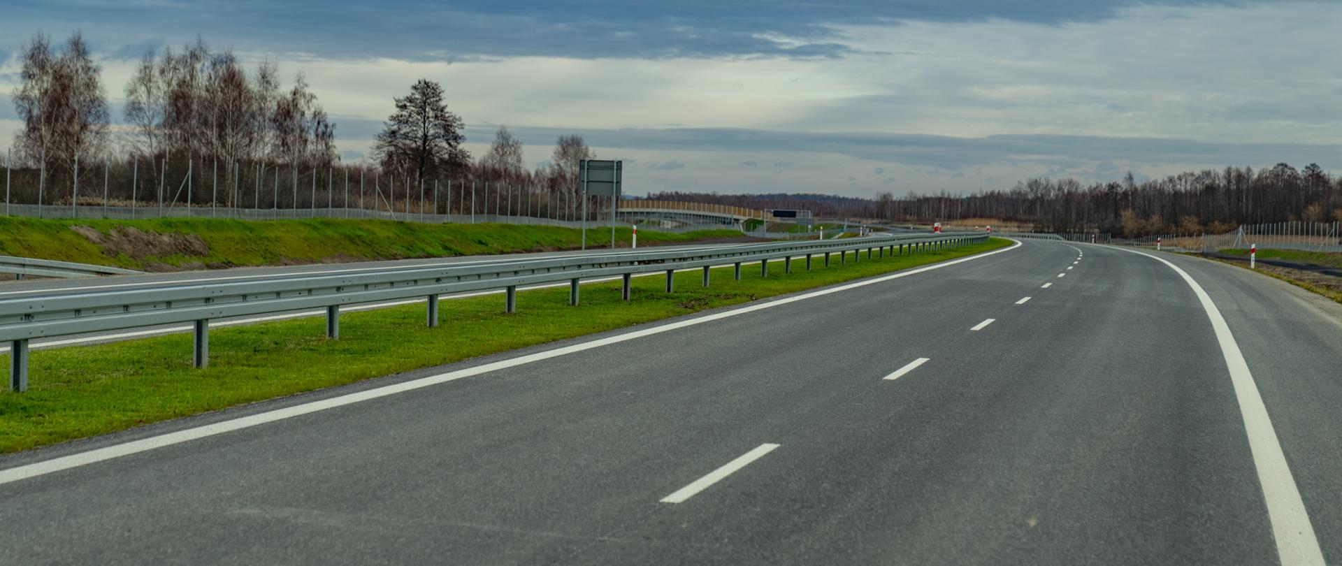 Na zdjęciu widać pustą, nową drogę ekspresową z dwoma pasami w każdym kierunku, oddzielonymi linią przerywaną. Po lewej stronie biegnie metalowa bariera ochronna i pas zielonej trawy, za którym znajduje się ogrodzenie oraz rząd drzew bez liści. Droga łagodnie skręca w prawo, a w tle widoczne są tablice informacyjne. Niebo jest częściowo zachmurzone w odcieniach szarości i błękitu.