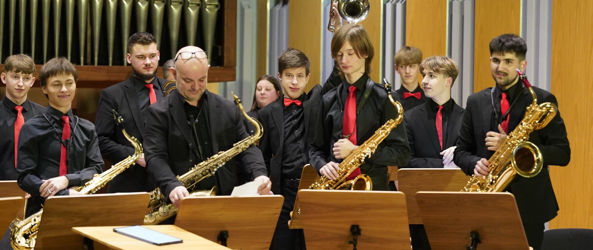Kilkuosobowy zespół młodych muzyków na scenie w sali koncertowej.
Wszyscy w ciemnych strojach, z czerwonymi krawatami; na pierwszym planie kilku gra na saksofonach, jeden muzyk podaje kartki. Za nimi widać rząd stojaków na nuty i fragment dużego instrumentu piszczałkowego, prawdopodobnie organów. Muzycy stoją lub siedzą, gotowi do grania, atmosfera formalna, jak podczas koncertu czy występu.