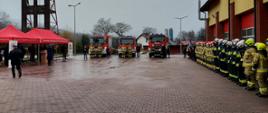 Zdjęcie przedstawia plac uroczystości. Po prawej stoją strażacy w dwuszeregu, natomiast po środku stoją trzy średnie samochody ratowniczo - gaśnicze. 