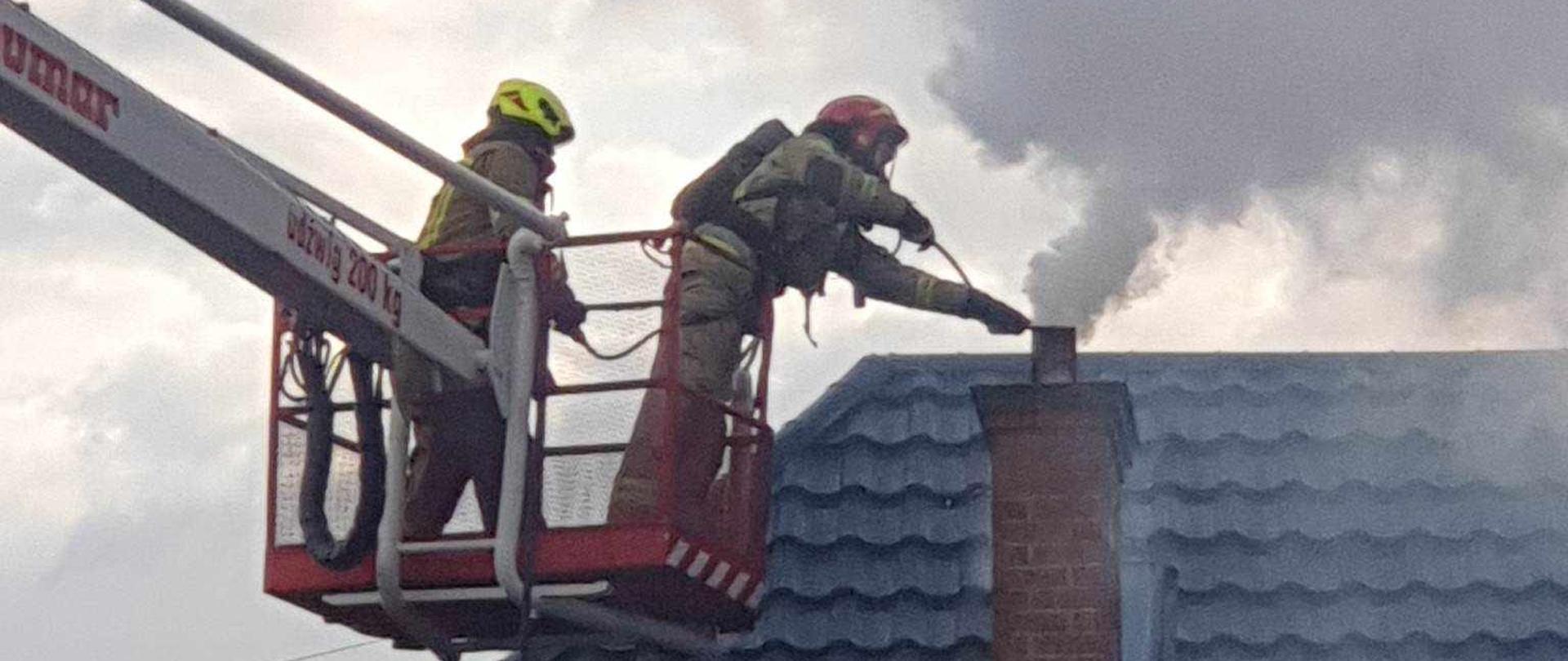 Zdjęcie przedstawia dwóch strażaków znajdujących się w koszu podnośnika. Kosz znajduje się w pobliżu dachu. Na dachu znajduje się komin z którego wydobywają się kłęby ciemnego dymu. Strażacy próbują ugasić pożar sadzy w kominie. Ubrani są w ubrania specjalne w jasnym kolorze. Na głowach mają hełmy w kolorze czerwonym oraz żółtym. Jeden ze strażaków na twarzy dodatkowo ma założoną maskę ochronną połączoną przewodem z butlą z powietrzem znajdującą się na jego plecach. 
