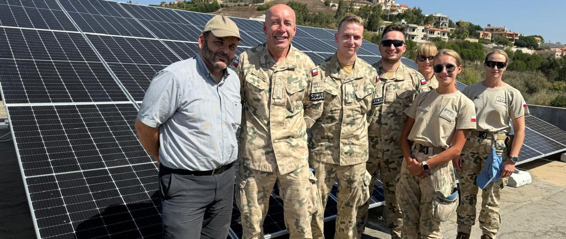 Trzech mężczyzn w mundurach w kamuflażu wojska polskiego z polskimi flagami i trzy kobiety w mundurach wojska polskiego w polskim kamuflażu z flagami pozuje na tle paneli słonecznych z cywilem 