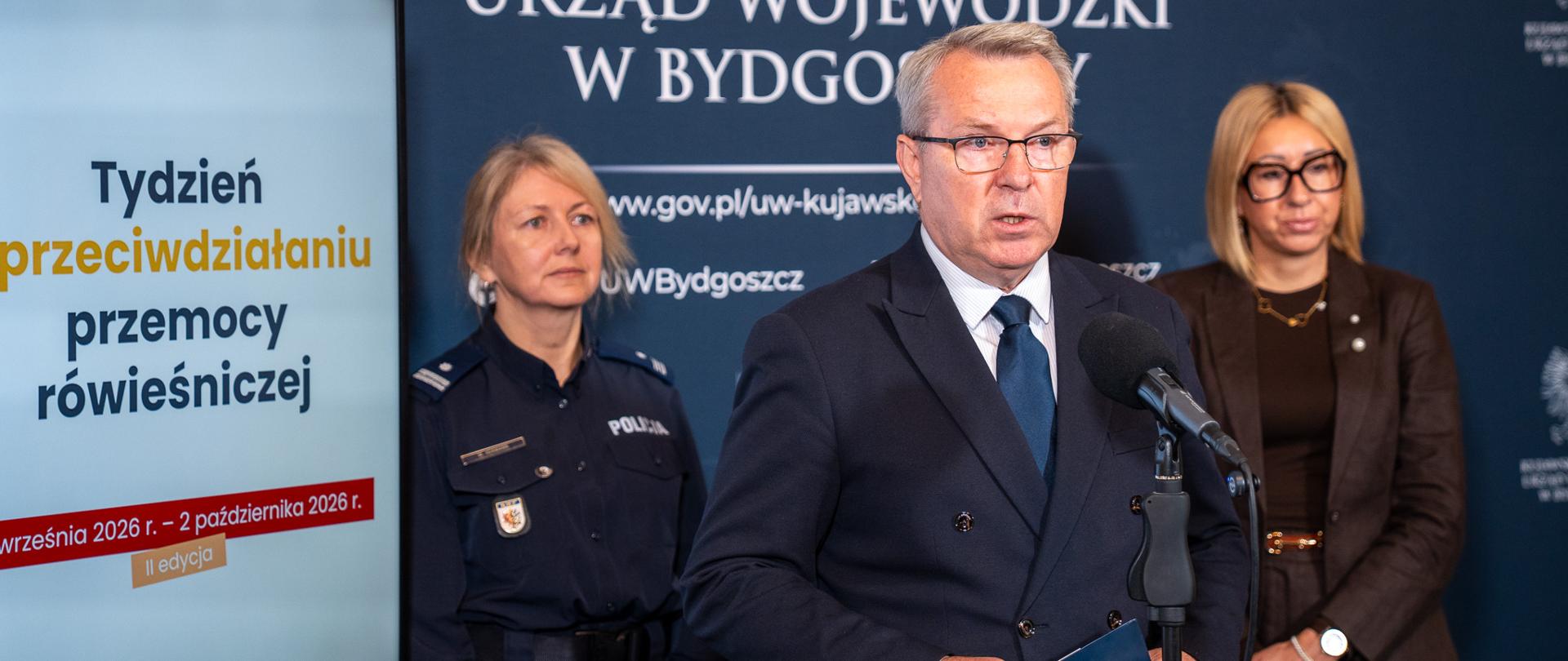 Wicewojewoda Piotr Hemmerling przemawia do mikrofonu, trzymając w rękach granatową teczkę z godłem Polski. Za nim stoją policjantka w mundurze oraz kobieta w okularach i brązowym żakiecie. Po lewej stronie widać ekran z napisem „Tydzień przeciwdziałania przemocy rówieśniczej”. 