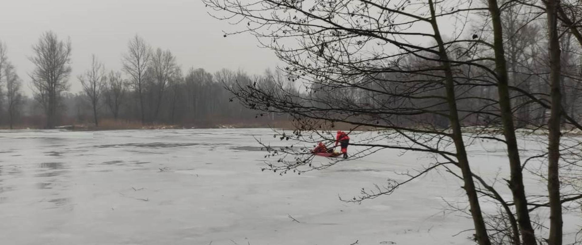 Dwóch strażaków wyposażonych w sanie lodowe podejmuje osobę poszkodowaną z wody. Działania na lodzie.