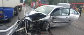 Zdjęcie przedstawia rozbity samochód osobowy marki Toyota Corolla na tle pojazdów strażackich oraz tira.
