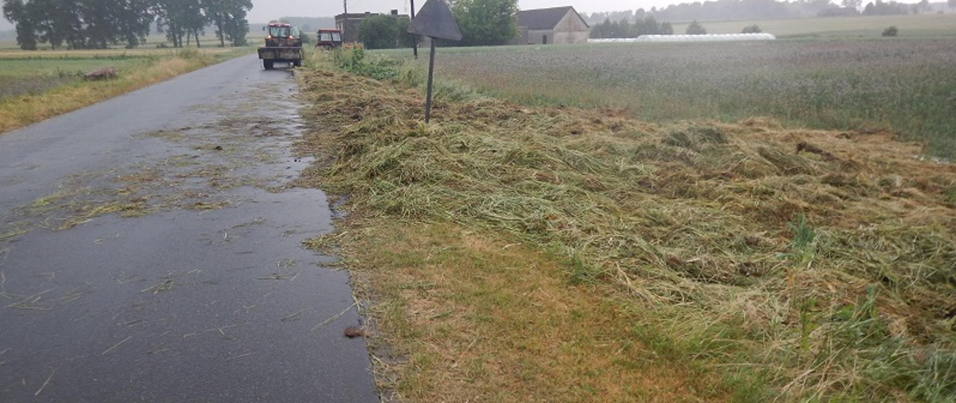 Zdjęcie przedstawia siano po pożarze na poboczu drogi. W oddali widoczne są 2 ciągniki rolnicze oraz przyczepa rolniczą.