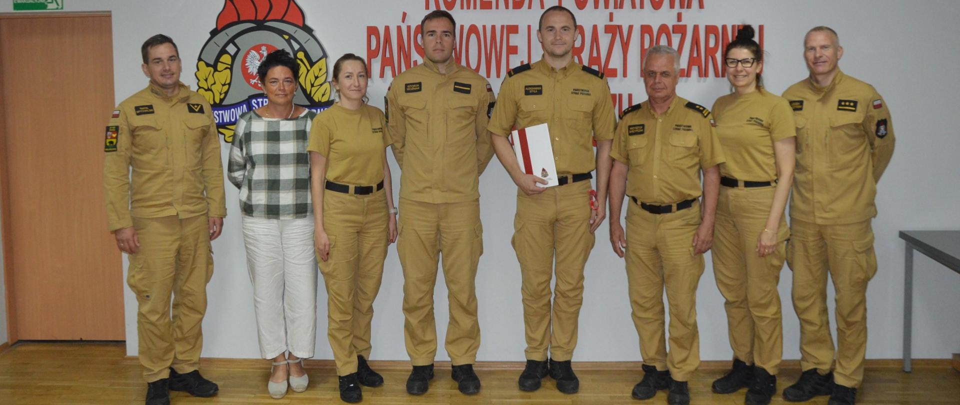 Świetlica Komendy Powiatowej PSP w Rawiczu. Na tle ściany, na której znajduje się czerwony napis: Komenda Powiatowa Państwowej Straży Pożarnej w Rawiczu stoi awansowany strażak w towarzystwie zastępcy komendanta powiatowego PSP oraz funkcjonariuszy i pracowników cywilnych komendy. Funkcjonariusze maja na sobie mundury służbowe w kolorze piaskowym. 