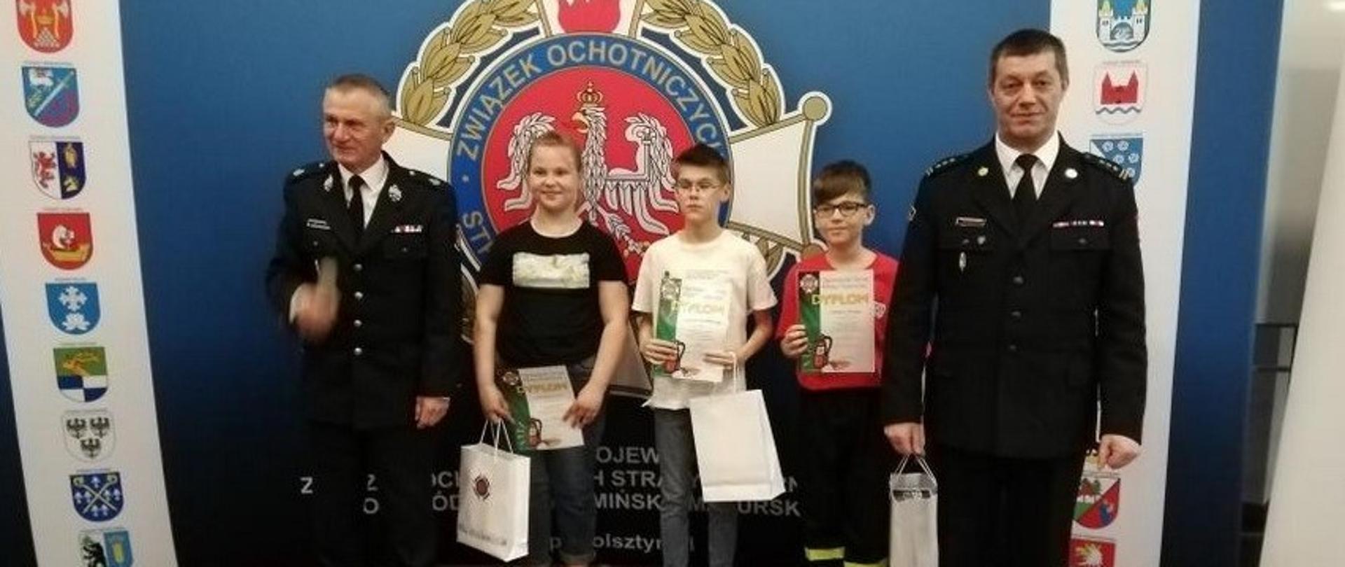 Zdjęcie przedstawia m.in. Katarzynę Turowicz ze Szkoły Podstawowej w Grabowie, gm. Gołdap, która zajęła II miejsce w swojej kategorii wiekowej (I grupa – szkoły podstawowe, klasy I–IV) podczas eliminacji wojewódzkich Ogólnopolskiego Turnieju Wiedzy Pożarniczej „Młodzież Zapobiega Pożarom”, które odbyły się w Bibliotece Uniwersyteckiej Uniwersytetu Warmińsko–Mazurskiego w Olsztynie. Na obrazie widać również przedstawicieli Komendy Wojewódzkiej Państwowej Straży Pożarnej w Olsztynie i Zarządu Oddziału Wojewódzkiego Związku Ochotniczych Straży Pożarnych RP woj. warmińsko-mazurskiego