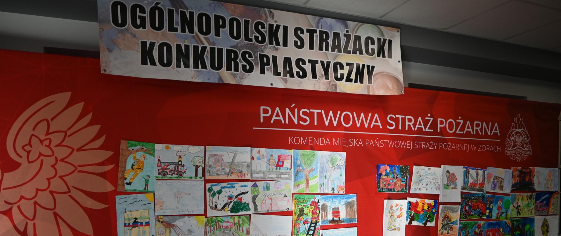 Na zdjęciu widać ekspozycję prac w ramach Ogólnopolskiego Strażackiego Konkursu Plastycznego. Kolorowe rysunki dzieci zostały wywieszone na dużej, czerwonej planszy z napisem „Państwowa Straż Pożarna – Komenda Miejska Państwowej Straży Pożarnej w Żorach"
