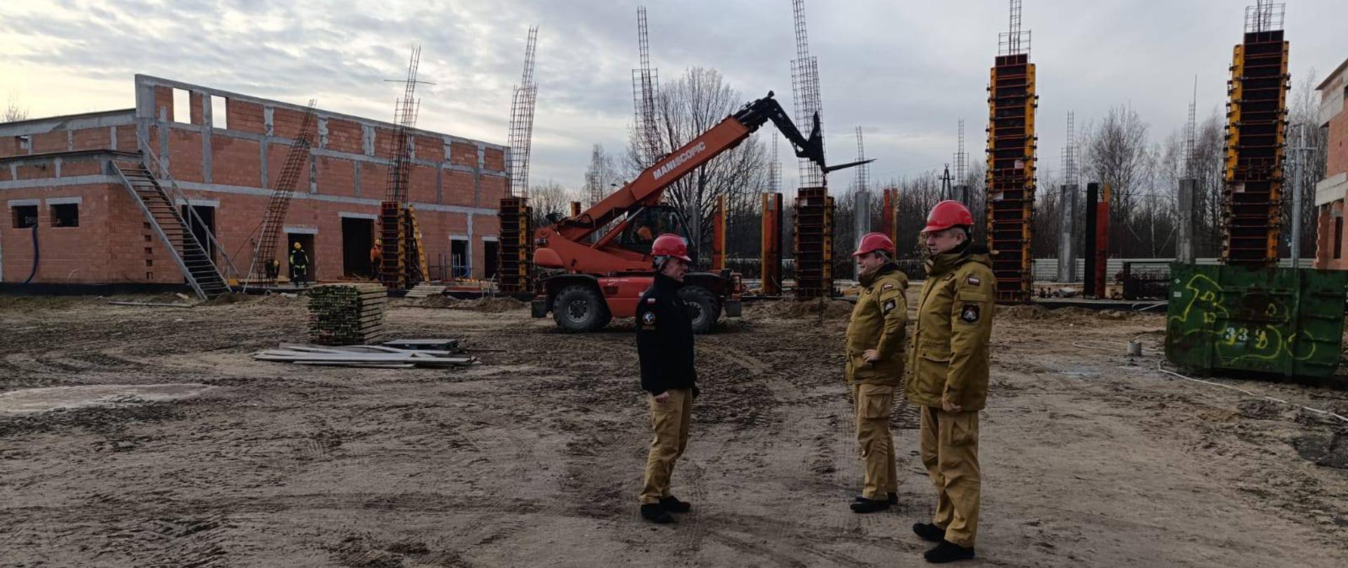 Trzech strażaków PSP w piaskowych i piaskowo-czarnych mundurach służbowych i czerwonych kaskach stoją na placu budowy. Za nimi po lewej stronie widoczny ceglasty budynek w budowie oraz liczne elementy konstrukcyjne. 