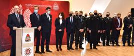 mężczyzna stoi przy mównicy i przemawia obok inne osoby w garniturach i kobieta dalej strażacy za nimi czerwona ścianka z logiem PSP