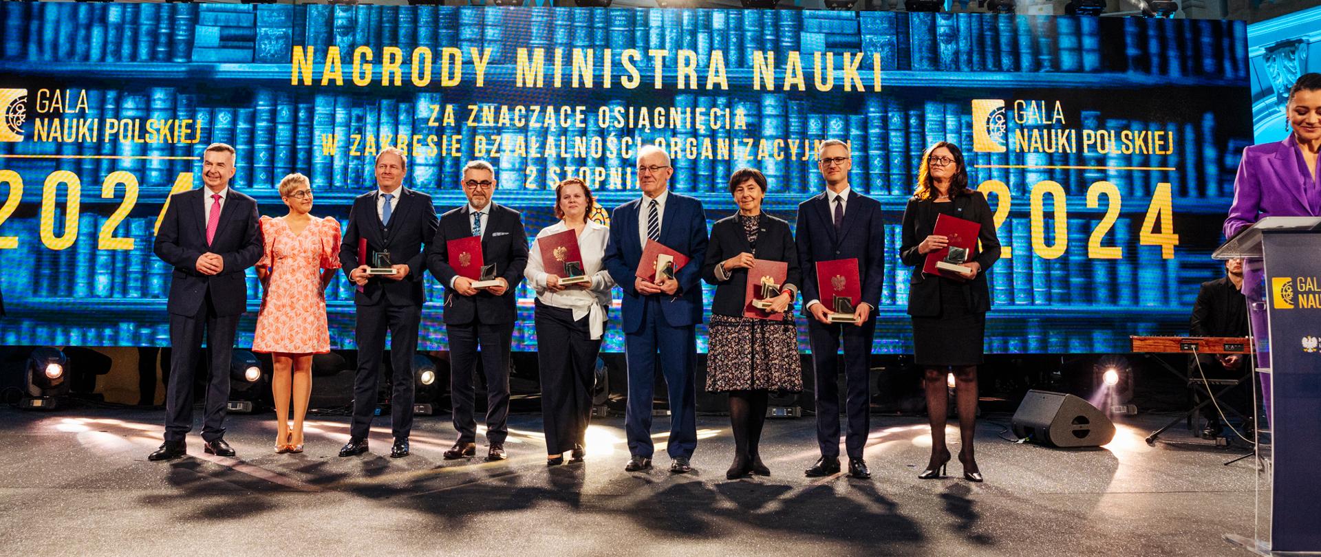 Na tle wielkiego ekranu z napisem Nagrody ministra stoi minister Wieczorek, wiceminister Mrówczyńska i kilka osób trzymających czerwone teczki.