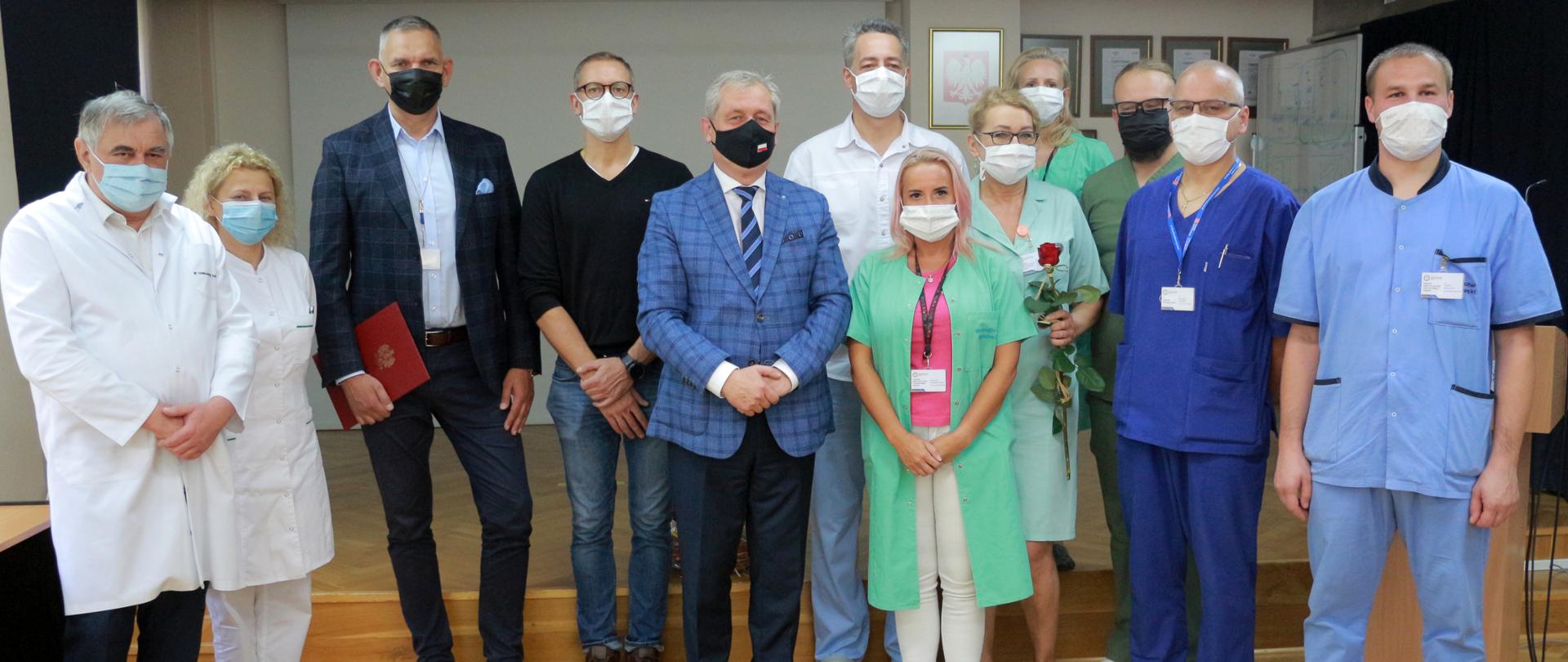 Na zdjęciu wojewoda opolski wraz z lekarzami, pielęgniarkami i dyrekcją Uniwersyteckiego Szpitala Klinicznego w Opolu. Łącznie na zdjęciu jest 11 osób. Wszyscy pozują do zdjęcia. Kadra medyczna ubrana jest w białe kitle. Wojewoda stoi po środku, w asyście pielęgniarki. Pozujący do zdjęcia stoją na tle ściany.