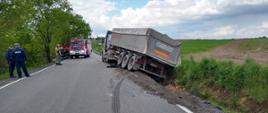 Prosty odcinek drogi asfaltowej. Po prawej stronie na skraju jezdni stoi ciągnik siodłowy z naczepą przechyloną silnie w prawą stronę. Koła naczepy wpadły do przydrożnego rowu. Przed naczepą na asfalcie widoczne ślady hamowania. Po lewej stronie jezdni stoi dwóch policjantów. W głębi na jezdni widoczny samochód straży pożarnej przed nim kilku strażaków oraz osobę cywilną