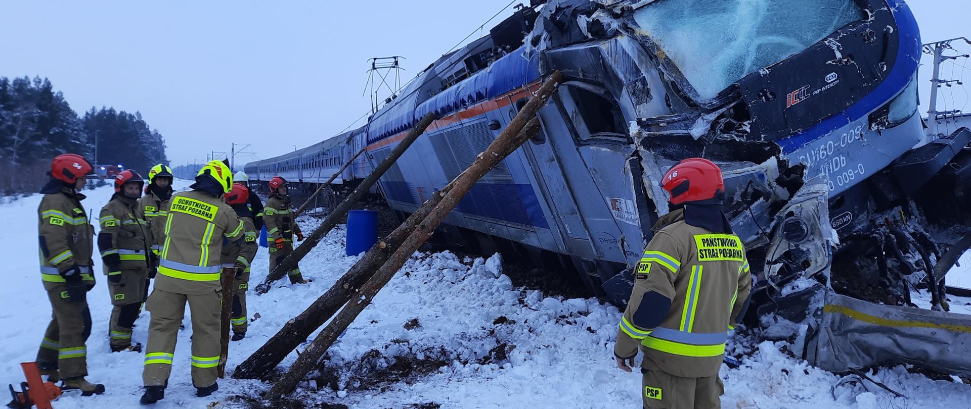 Na zdjęciu przechylona na bok lokomotywa wykolejona z boku podparta drewnianymi słupami. Wokoło strażacy PSP i OSP. Dookoła leży śnieg.
