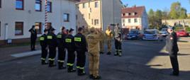 Uroczystość z okazji Dnia Flagi Rzeczypospolitej Polskiej. Strażacy ze zmian służbowych stoją naprzeciwko siebie. Poczet flagowy wciąga flagę państwową na maszt. Komendant, poczet flagowy dowódcy zmian salutują.