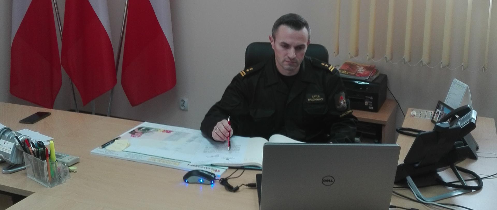 Zdjęcie Komendanta Powiatowego w Busku-Zdroju w gabinecie podczas wideokonferencji. w tle flagi Polski.