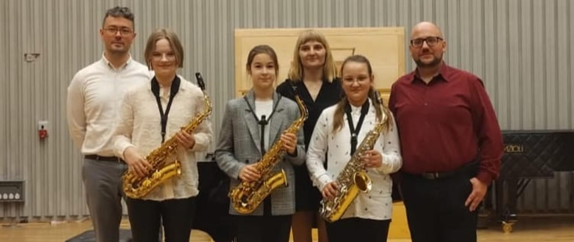 Zdjęcie przedstawia stojących na scenie trzymających saksofony trzech uczniów biorących udział w EFS wraz z nauczycielem Adrianem Masselim i akompaniatorami - naucz. Marią i Danielem Rybakami