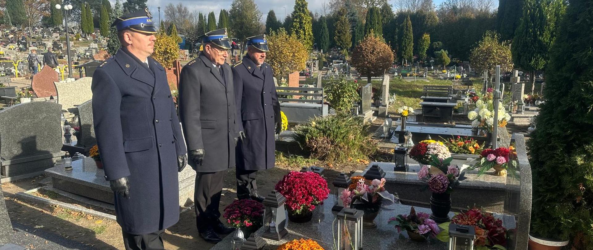Na zdjęciu widać trzech mężczyzn w mundurach galowych, stojących na cmentarzu przy jednym z grobów. Mają na sobie ciemne płaszcze, czapki z daszkiem i czarne rękawiczki. Na grobie leży wiele kolorowych kwiatów – głównie chryzantem – oraz kilka zniczy. W tle widać liczne inne nagrobki, drzewa i krzewy, a nad nimi częściowo zachmurzone niebo. Scena wygląda na moment zadumy i oddania hołdu z okazji Święta Wszystkich Świętych