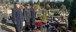 Na zdjęciu widać trzech mężczyzn w mundurach galowych, stojących na cmentarzu przy jednym z grobów. Mają na sobie ciemne płaszcze, czapki z daszkiem i czarne rękawiczki. Na grobie leży wiele kolorowych kwiatów – głównie chryzantem – oraz kilka zniczy. W tle widać liczne inne nagrobki, drzewa i krzewy, a nad nimi częściowo zachmurzone niebo. Scena wygląda na moment zadumy i oddania hołdu z okazji Święta Wszystkich Świętych