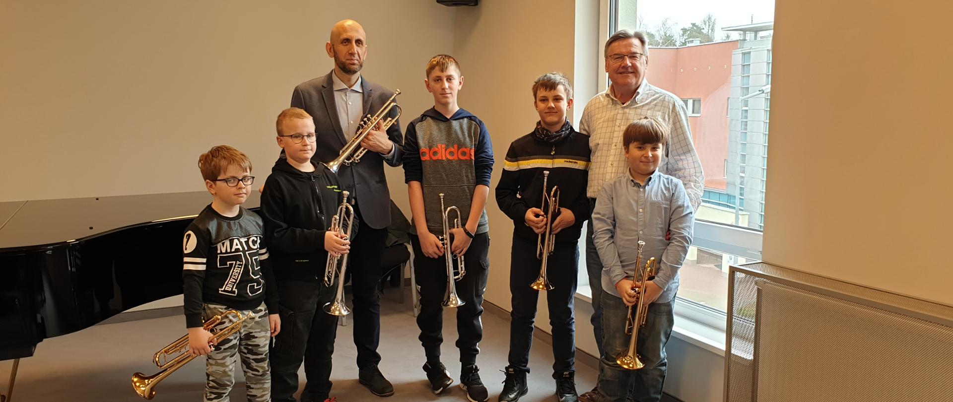 Zdjęcie na sali kameralnej uczniów wraz z nauczycielem i prowadzącym warsztaty. Uczniowie oraz prowadzący warsztaty trzymają w ręku trąbki, za nimi z prawej strony okno, z prawej strony fortepian