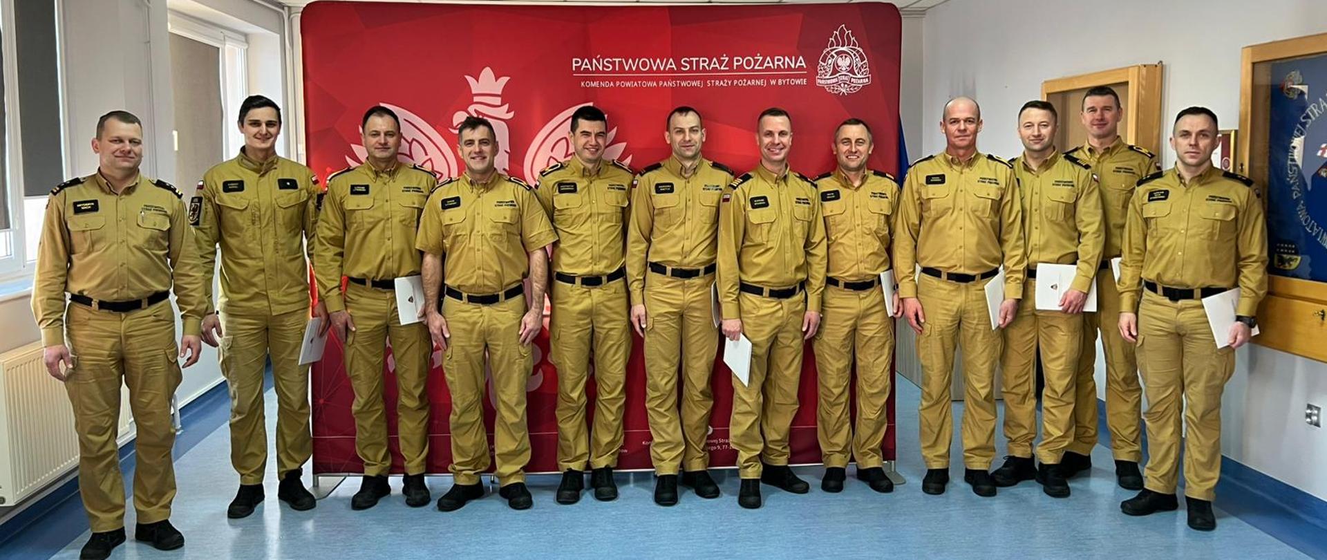 Wyróżnieni funkcjonariusze w szeregu pozują do zdjęcia na tle czerwonego baneru Państwowa Straż Pożarna