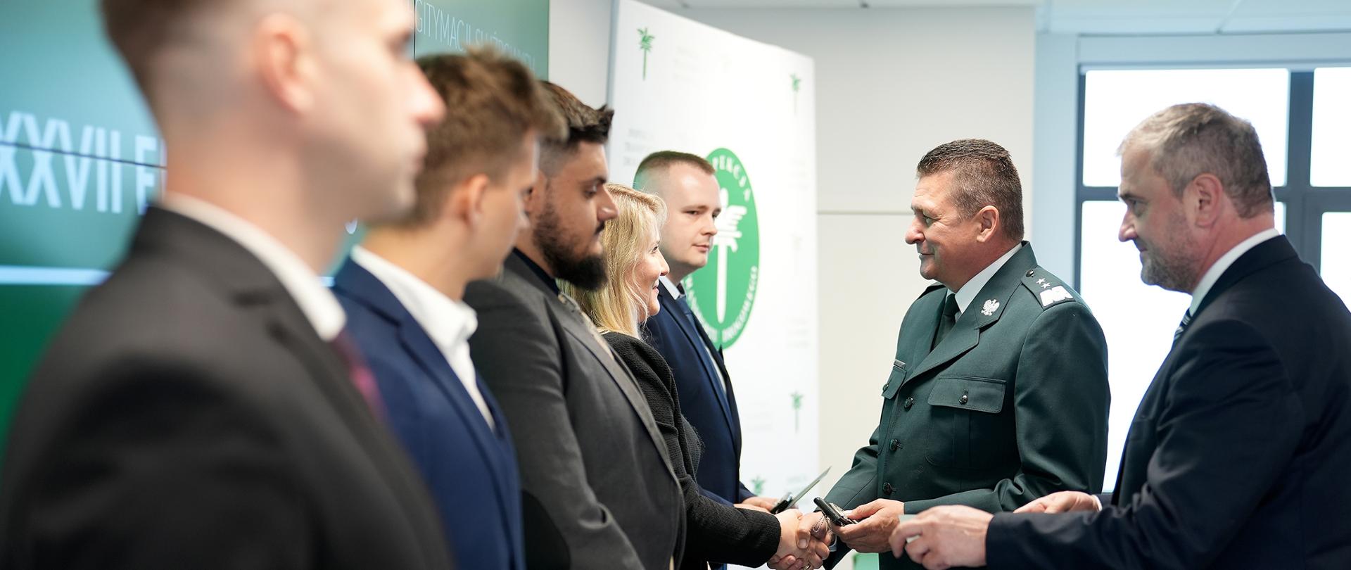 Mężczyzna w mundurze podaje rękę i legitymację służbową kobiecie stojącej wśród innych mężczyzn. Obok stoi mężczyzna w garniturze. 