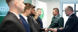 Mężczyzna w mundurze podaje rękę i legitymację służbową kobiecie stojącej wśród innych mężczyzn. Obok stoi mężczyzna w garniturze. 
