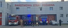 Na zdjęciu widać budynek straży pożarnej z dużym napisem „PAŃSTWOWA STRAŻ POŻARNA” nad wjazdami do garaży. Przed budynkiem stoją trzy pojazdy: dwa duże wozy strażackie z włączonymi niebieskimi światłami oraz mniejszy samochód osobowy, policyjny. Na tle budynku i pojazdów ustawiona jest grupa strażaków w mundurach, stojących w szeregu, oddając hołd zmarłemu strażakowi ochotnikowi z Ochotniczej Straży Pożarnej w Sierakowicach, który zginął 13 sierpnia 2025 roku w trakcie prowadzonych działań ratowniczo-gaśniczych podczas pożaru zakładu produkcyjnego w Kawlach . Po lewej stronie stoi strażak – dowódca ubrany w mundur służbowy koloru beżowego. Pozostali strażacy siedem osób ubranych w ubrania strażackie koloru czarnego. Po lewej stronie stoi policjant ubrany w mundur policyjny koloru granatowego. Cała scena odbywa się na placu z kostki brukowej, przy jasnym niebie z lekkim zachmurzeniem