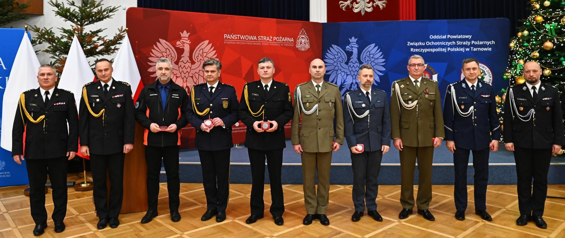 W rzędzie przed sceną stoją uczestnicy gali funkcjonariusze ubrani są mundury galowe. Obok sceny znajdują się choinki. Na ścianie wisi godło Polski, a na scenie znajdują się bilbordy czerwony Pastowej Straży Pożarnej, oraz niebieski Związku Ochotniczych Straży Pożarnych
