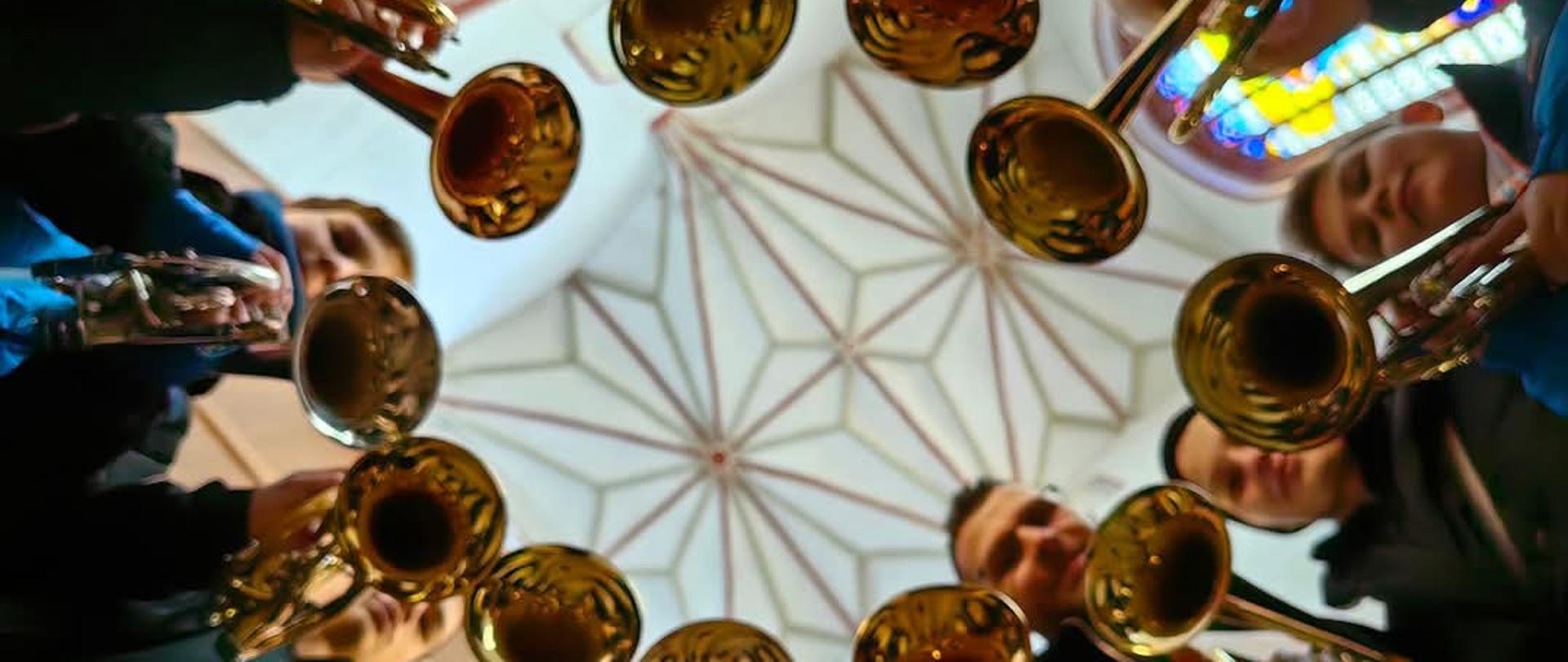Zdjęcie przedstawia zespół trąbek stojący w kole i trzymający instrumenty na tle gwiaździstego sklepienia kolegiaty Najświętszej Marii Panny Królowej Świata w Stargardzie.
