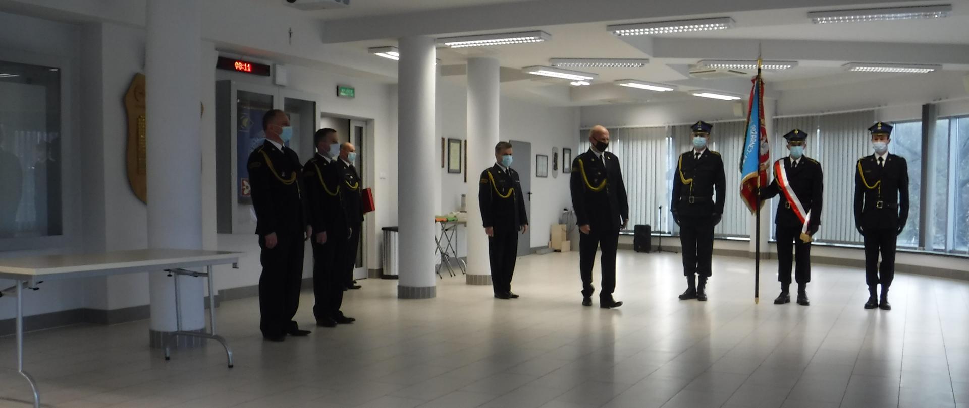 Na zdjęciu widzimy kilku strażaków podczas uroczystej zbiórki z okazji pożegnania strażaków przechodzących na zaopatrzenie emerytalne. Trzech strażaków stoi w postawie baczność w poczcie sztandarowym, jeden z nich trzyma sztandar. Strażacy ubrani są w mundury wyjściowe ze sznurem.