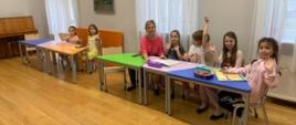 W klasie, przy kolorowych stolikach zasiadają dzieci oraz ich nauczycielka. Wszyscy uśmiechają sie w kierunku obiektywu.