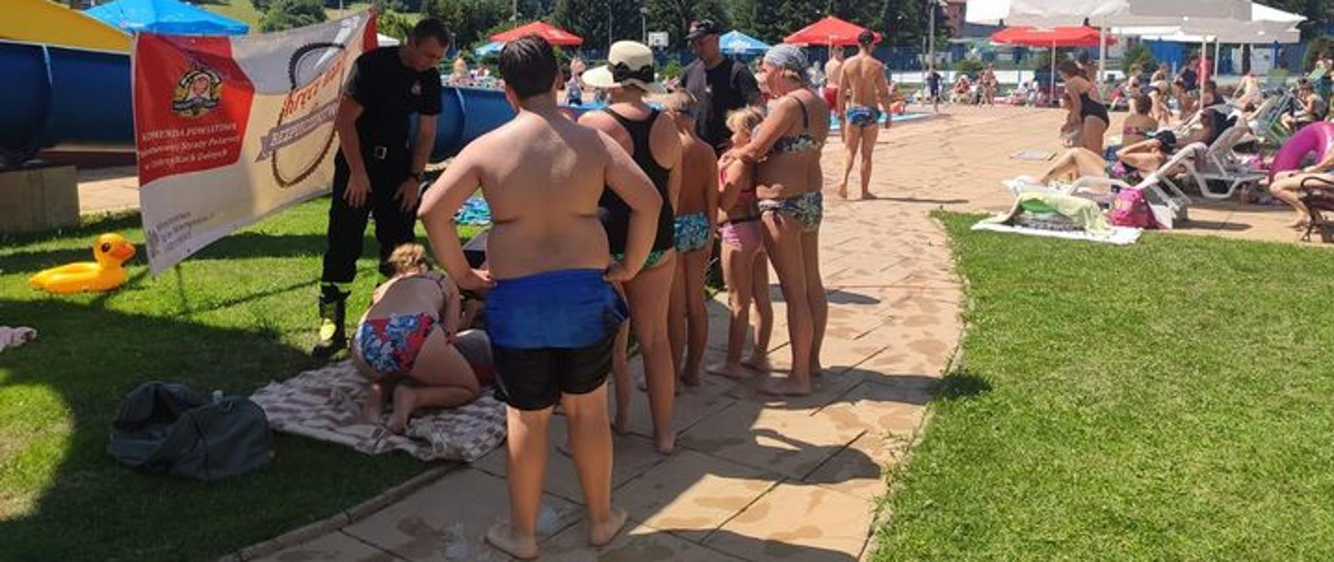 Na zdjęciu widać strażaków w ubraniach koszarowych którzy znajdują się na basenie. Pokazują Oni jak prawidłowo wykonać resustytacje krążeniowo-oddechową zgromadzonym przed nimi osobom. Za plecami jednego ze strażaków jest plakat pn. "Kręci mnie bezpieczeństwo" zawieszony na zjeżdżalni. W oddali znajdują się leżaki i parasole oraz ludzie. 