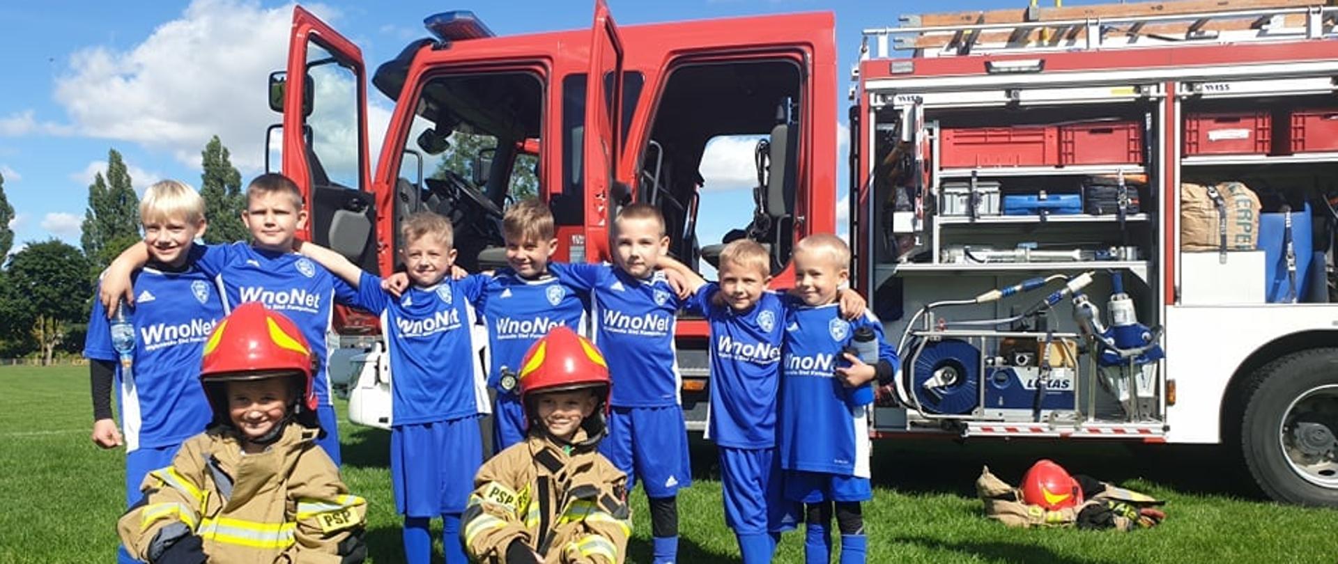Na zdjęciu widać grupę chłopców w strojach sportowych, którzy trzymają się za szyje. Przed nimi klęka dwóch chłopców w strażackich ubraniach specjalnych z czerwono-żółtymi hełmami na głowach. W tle widać wóz strażacki.