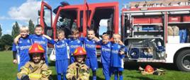 Na zdjęciu widać grupę chłopców w strojach sportowych, którzy trzymają się za szyje. Przed nimi klęka dwóch chłopców w strażackich ubraniach specjalnych z czerwono-żółtymi hełmami na głowach. W tle widać wóz strażacki.