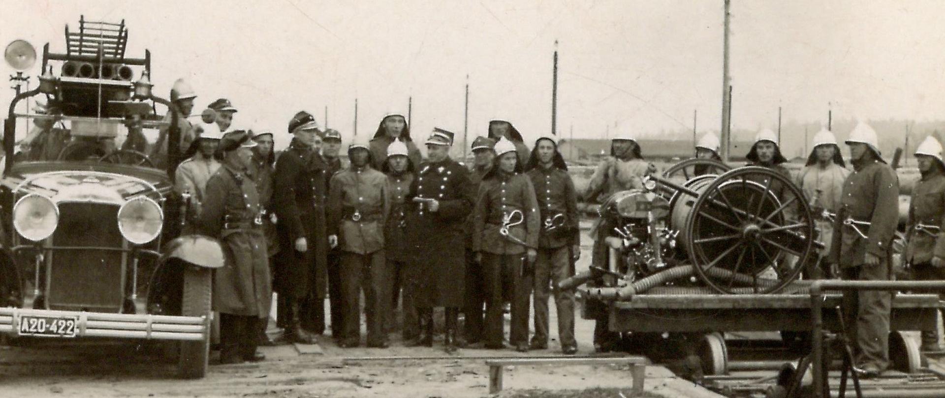 Na tle wozu strażackiego stoi kilkunastu strażaków w mundurach