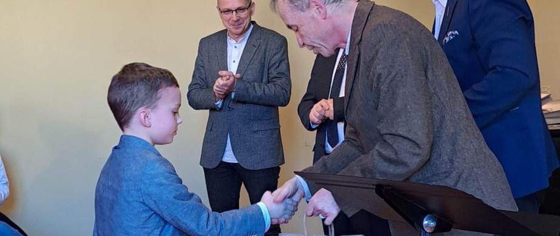 Zdjęcie przedstawia ucznia Miłosza Matyszczaka odbierającego nagrodę ogłoszenia wyników Marcowego Konkursu Klarnetowego 2023 w Szczecinie.