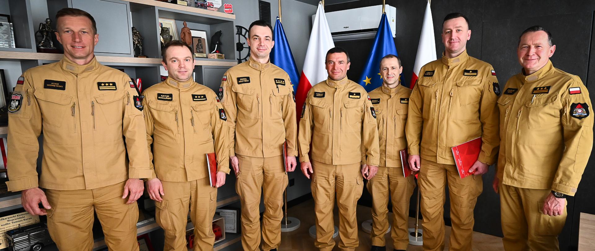 Wspólne zdjęcie pamiątkowe siedmiu oficerów PSP w piaskowych mundurach. Stoją w jednym rzędzie na tle flag i regału z pucharami. Od lewej: st. bryg. Adam Wiśniewski, kpt. Artur Barański, st. kpt. Paweł Krzyśko, nadbryg. Tomasz Baran, mł. bryg. Marek Rusnarczyk, mł. bryg. Tomasz Pustelak oraz st. bryg. Dariusz Homa.