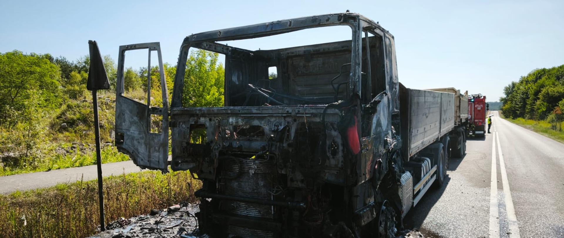 Na zdjęciu widać spaloną kabinę ciężarówki stojącą na poboczu asfaltowej drogi, z otwartymi drzwiami kierowcy. Przód pojazdu jest całkowicie zniszczony przez ogień, a wokół widoczna jest spalona i osmolona ziemia. Za ciężarówką stoi nienaruszona część naczepy oraz czerwony wóz strażacki, a w tle widać prosty odcinek drogi i zielone zadrzewienia po obu stronach.