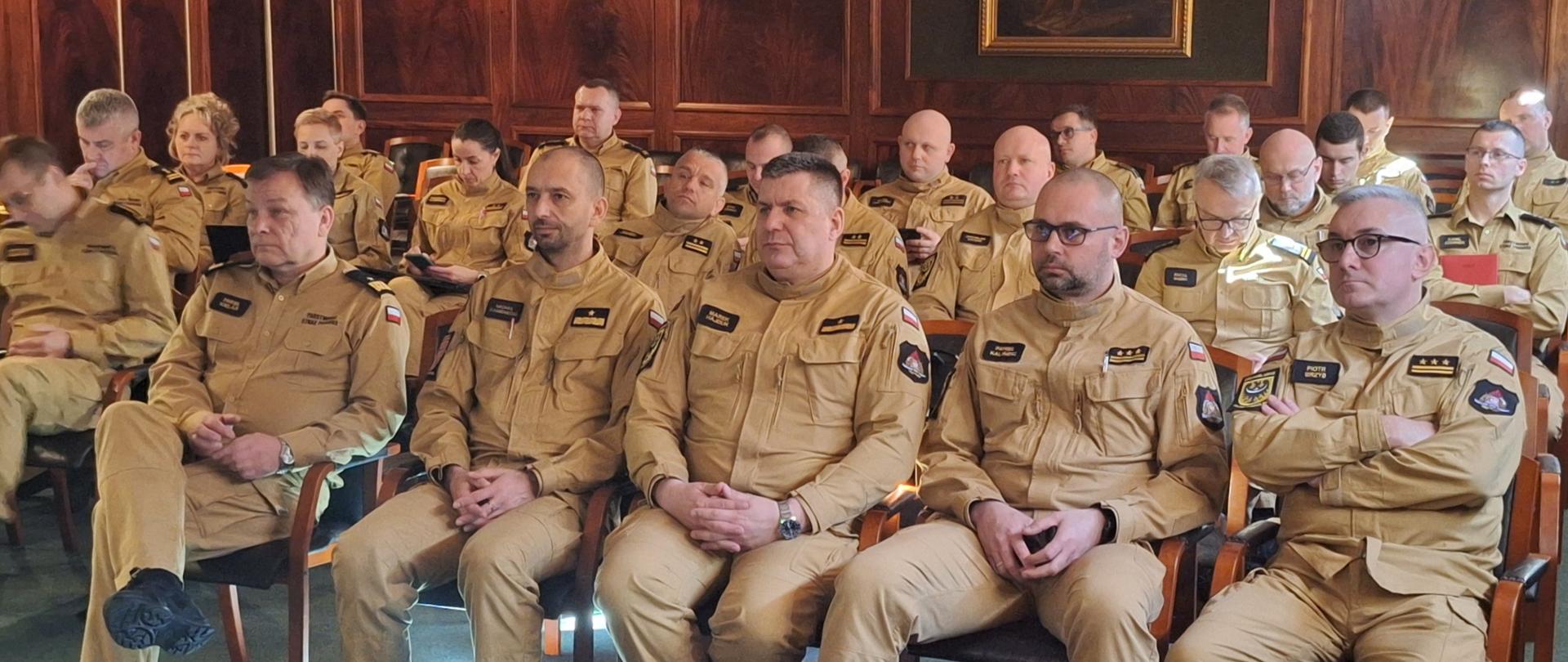 Funkcjonariusze Państwowej Straży Pożarnej podczas warsztatów tematycznych poświęconych analizie działań ratowniczych oraz zarządzania kryzysowego podczas i po powodzi. Strażacy w umundurowaniu służbowym słuchają wykładu funkcjonariusza Państwowej Straży Pożarnej. Zdjęcie na sali wykładowej.