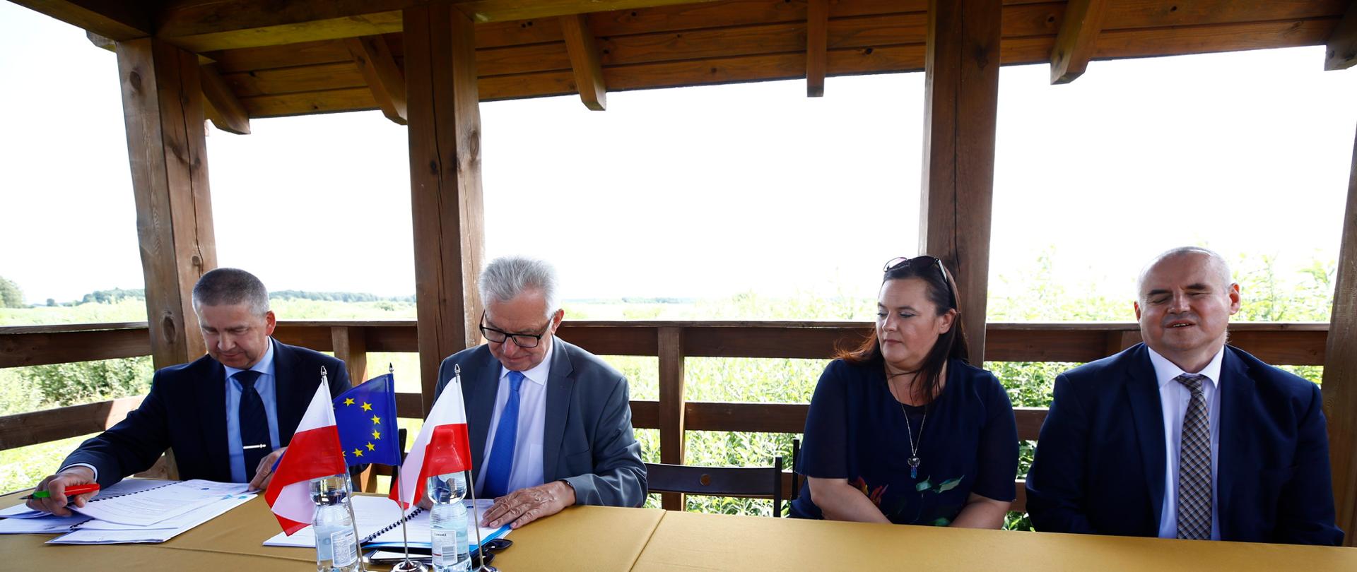 przy stole siedzą dwie podpisujące umowę osoby przed którymi stoją proporczyki z flagami Polski i Unii Europejskiej, obok przy stole dwoje wiceministrów, w tle zielona łąka