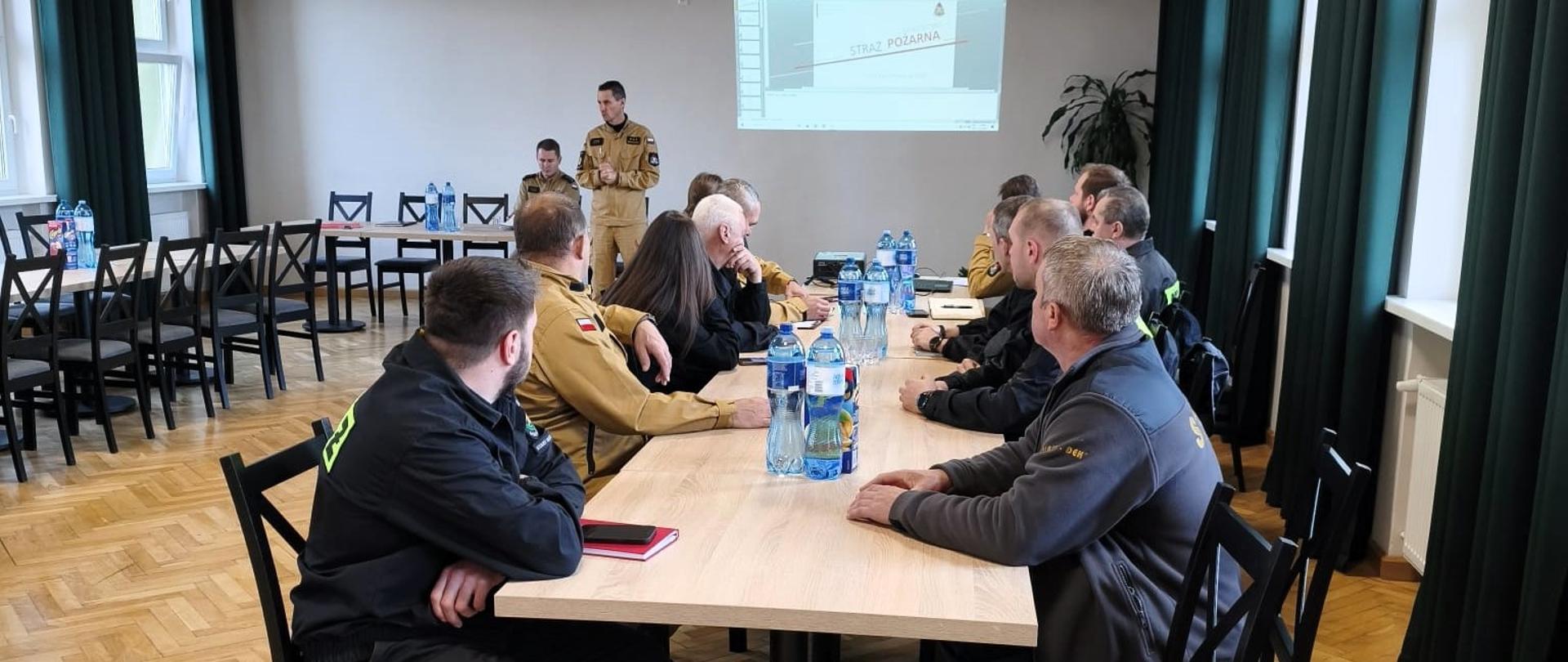 W jasnej sali szkoleniowej kilku strażaków siedzi przy prostokątnym stole, słuchając prowadzącego. Prowadzący stoi z przodu sali w ubraniu koszarowym koloru beżowego i prezentuje treści wyświetlane na ścianie z projektora. Uczestnicy siedzą tyłem lub bokiem do aparatu, na ich kurtkach widoczny jest napis „STRAŻ”. Na stole stoją butelki z wodą i notatniki. Sala jest przestronna, z drewnianą podłogą i dużymi oknami po prawej stronie.
