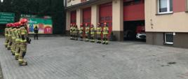 Przed budynkiem komendy PSP w Mławie stoją strażacy w dwóch szeregach na przeciwko siebie. Pomiędzy nimi stoi dowódca JRG Mława. Jedna brama garażu jest uchylona, widać pojazd pożarniczy.