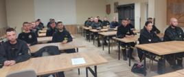 Strażacy ubrani w czarne ubrania koszarowe siedzą w ławkach przodem skierowani do stołu prezydialnego. Z tyłu widać ścianę z ekranem do wyświetlania prezentacji