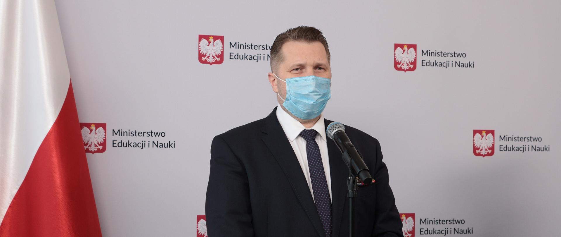 Minister Przemysław Czarnek stoi przy mikrofonie. Na twarzy ma maseczkę, jest ubrany w garnitur. W tle baner z logotypami Ministerstwa Edukacji i Nauki i biało-czerwona flaga. 